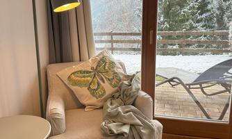 A cozy armchair with a blanket and a pillow is next to a small table. Through the window, you can see snow-covered trees and a balcony.