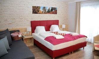 A modern bedroom with a red bed and a cozy couch. The room has large windows and a bright, inviting look.