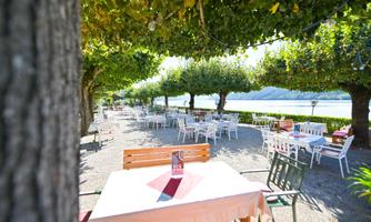 An inviting restaurant garden by the water with many tables under shady trees. The place is bright and friendly, ideal for a relaxed meal outdoors.