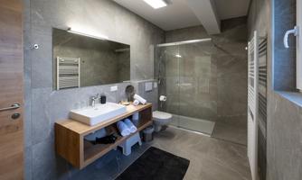 A modern bathroom with elegant tiles and natural wood elements. It features a spacious shower, a large sink, and practical storage spaces.