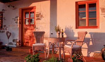 A cozy porch with two chairs and a table. On the table, there are wine and a plant, while the evening sun shines on the facade.
