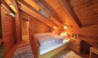 A cozy bedroom with wooden walls and a skylight. It contains a double bed, a bedside table, and a colorful carpet.
