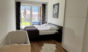 A bright bedroom with a large window overlooking the garden. There is a bed, a crib, and a rug on a wooden floor.