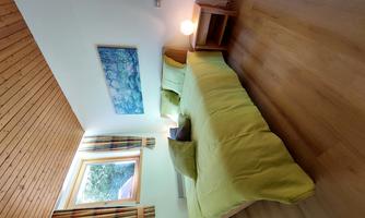 A bright bedroom with a double bed in green bedding and a large window. The walls are white and the wooden ceiling creates a cozy atmosphere.
