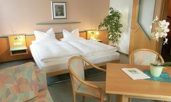 A cozy hotel room with two beds, a table and chairs. The decor is bright and friendly, with plants for decoration.