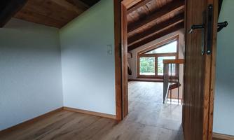 A bright room with wooden floors and slanted ceilings. In the background, there is a window that offers a view of greenery.