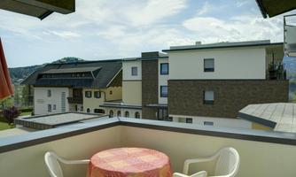 A balcony with a round table and plastic chairs. In the background, several modern buildings and green hills can be seen.