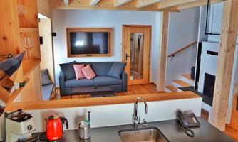 A modern living-dining room with a gray sofa and a wood stove. In the background, a modern kitchen and a staircase can be seen.