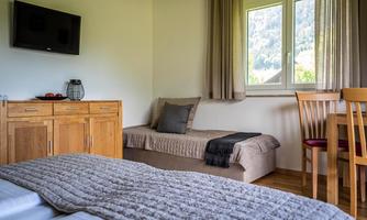 A cozy room with two beds, a wooden wardrobe, and a small table. Bright daylight streams through the window with a view of nature.