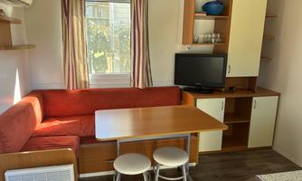 A cozy living area with a red sofa and a wooden table. A TV stands on the wall and there are large windows with curtains.