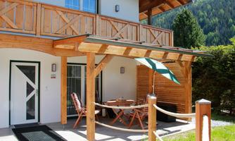 A cozy terrace with a wooden veranda and two lounge chairs. In the background, green trees and a clear sky can be seen.