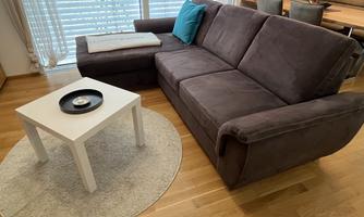 A modern living room with a comfortable gray sofa and a small white table. The room has large windows with a view outside and a bright, inviting floor.
