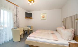 A bright room with two single beds, a television, and a small table with chairs. Large windows let in plenty of natural light.