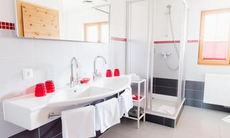 A modern bathroom with a shower cabin and a double sink. Red towels and a reddish bath mat give the room a fresh accent.