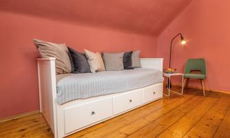 A cozy room with a white daybed and cushions in soft colors. Next to the bed is a small table with a lamp and a green chair.
