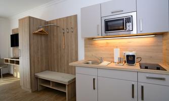 A modern kitchenette with wooden fronts and a microwave oven. In the background, there is a wooden shelf with a hook and a bench.