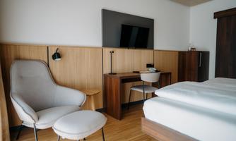 A modern hotel room with a cozy armchair, a desk, and a flat-screen TV on the wall. The warm wood tones create an inviting atmosphere.