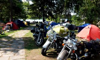 A campsite with several tents and parked motorcycles. Between the tents, trees and a clearing can be seen.