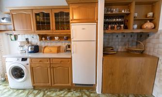 A modern kitchen with wooden cabinets and a washing machine. The refrigerator is next to a wooden shelf with spices and decoration.
