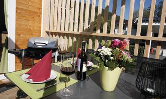 A cozy table on a balcony with a BBQ grill, red wine, and flowers. The scene is bright and inviting under a blue sky.
