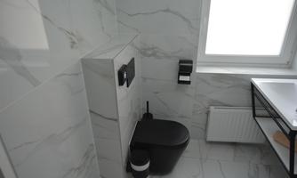 A modern bathroom with simple, gray tiles. The toilet is elegant and black, next to it is a sink with a minimalist design.