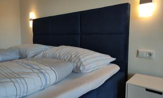 A modern bedroom with an elegant blue headboard and white bedding. Next to the bed is a minimalist nightstand with soft lighting.
