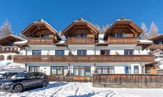 A cozy building in the snow with wooden balconies and a clear blue sky. In front of the house, there is a black car.