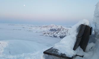 A snow-covered mountain landscape with an impressive view of clouds and mountains. The sky is softly tinted, and the moon is visible.