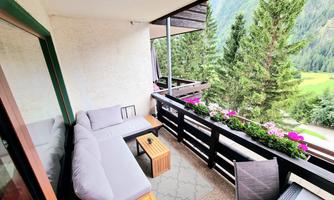 A cozy balcony with a seating area and a small table. In the background, tall trees and a beautiful landscape can be seen.
