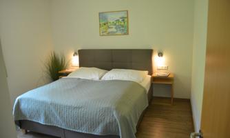 A cozy bedroom with a large double bed and bright bedding. A colorful painting hangs on the wall, and there is a pleasant plant arrangement.