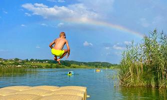 A boy jumps from a dock into a clear lake. In the background, there is a rainbow and the landscape is green and hilly.