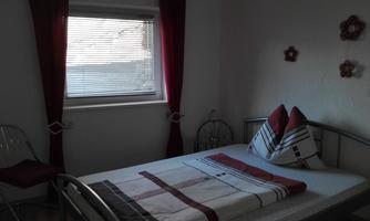 A simple bedroom with a bed and colorful pillows. There is a chair and red curtains at a window.
