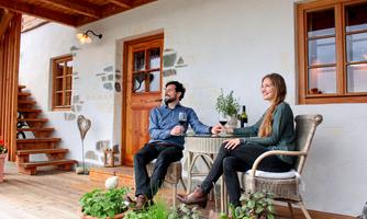 A cozy outdoor area with a round table, where a man and a woman are sitting. In the background, there is a wooden house and some plants visible.
