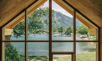 A beautiful view from a modern wooden house overlooking a tranquil lake surrounded by trees and mountains. The landscape radiates tranquility and connection to nature.