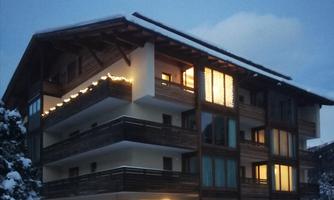 A modern wooden house in winter, surrounded by snow. The balconies are decorated with lights and warm light radiates from the windows.