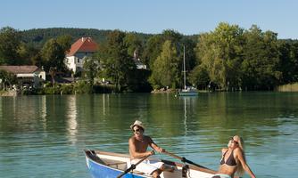 A boat on a calm lake, surrounded by trees and houses. Two people are relaxing in the boat in sunny weather.