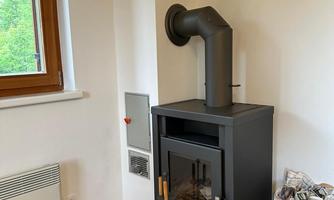 A modern wood stove in a bright corner of a room. Next to it are firewood logs and a radiator.