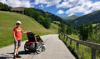 A boy stands at the roadside in a picturesque mountain landscape. Next to him is a stroller on the path.