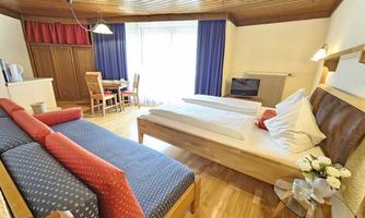 A cozy hotel room with two beds, a sofa, and a small dining table. Large windows provide plenty of daylight and an inviting atmosphere.