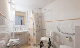 A bright bathroom with a shower, a sink, and a toilet area. The furnishings are accessible for people with disabilities, featuring grab bars and a shower chair.