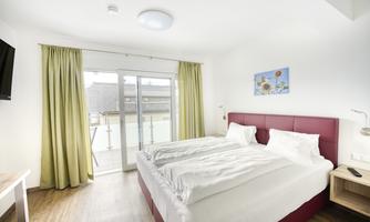 A modern bedroom with a large double bed and white bed linens. Green curtains hang in front of the windows, and there is access to a balcony.