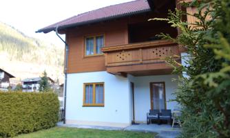 A beautiful wooden house with a balcony and a well-maintained garden. In the background, mountains and a green landscape can be seen.
