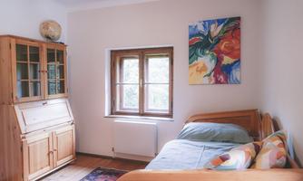 A cozy bedroom with a single bed and colorful cushions. A colorful painting hangs on the wall and there is a wooden desk with a window that lets in daylight.