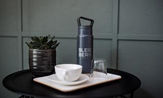 A stylish arrangement on a table with a water bottle, a cup, and a glass. In the background, there is a small plant in a black pot.