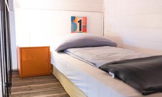 A simple bedroom with a bed, a nightstand, and minimalist decor. The walls are white and the floor is made of wooden planks.