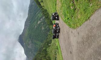 Two quads are standing on a narrow path in a green, mountainous landscape. The sky is cloudy and the surroundings are rich in nature.