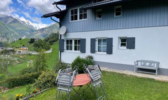 A cozy house in the mountains with a well-kept garden. In the foreground, there is a table with chairs, and the view shows green meadows and mountains.