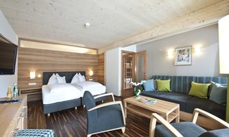 A modern room with two beds, a cozy sitting area, and wooden paneling. The room is bright and inviting, with decorative cushions and a TV.