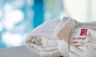 A white bathrobe with the "Moserhof" logo is lying on a table. In the background, a blue pool is visible.