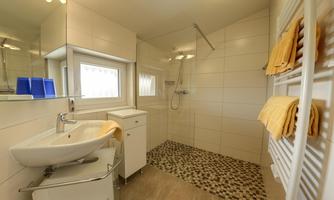 A modern bathroom with a shower, a washbasin, and a large mirror. The walls are light, and there are towels in soft colors.
